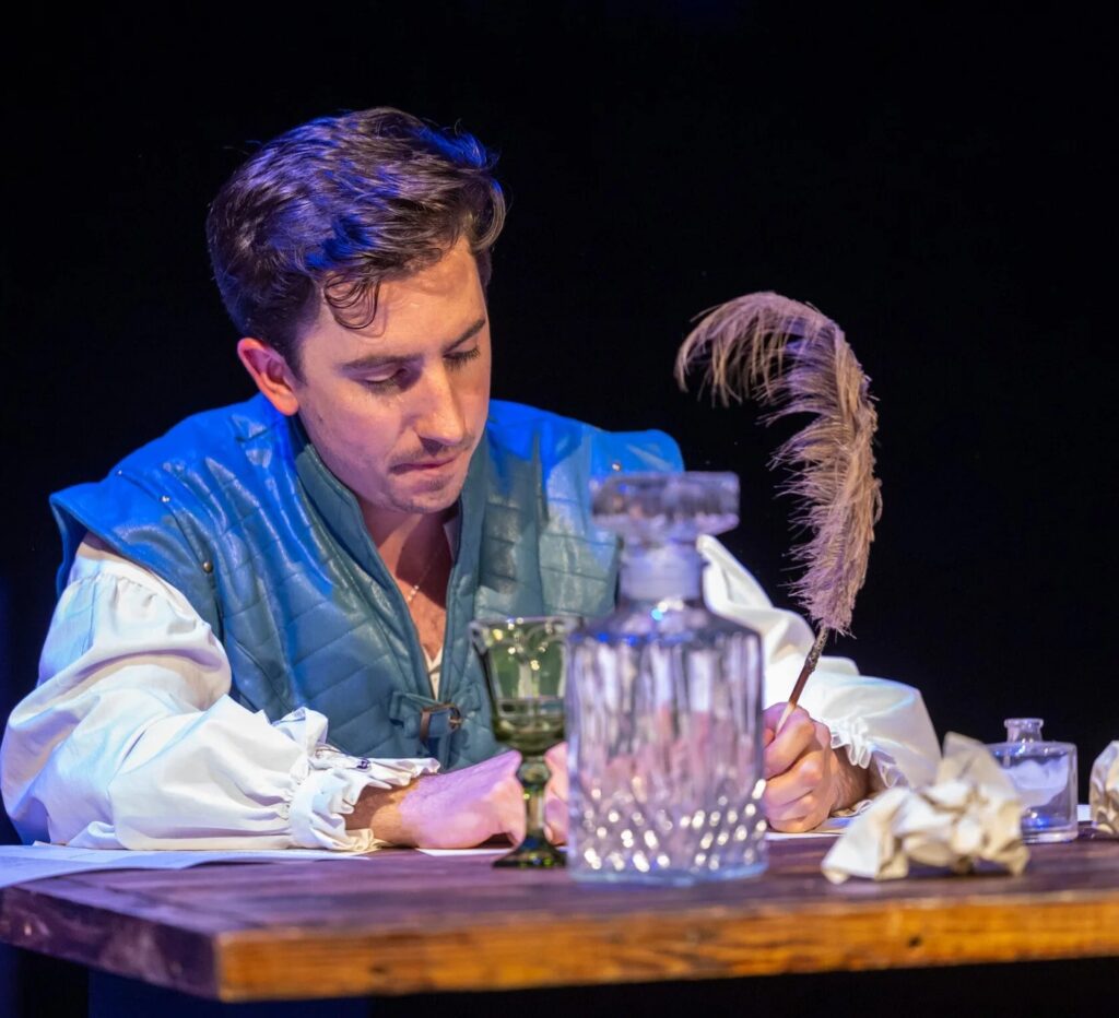 A performer in blue period costume writing at a candlelit desk